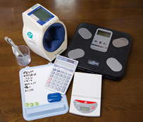 牧野さんの自己管理七つ道具。右上から時計回りに、体重計、はかり、電卓、ホワイトボード、計量カップとスプーン、血圧計